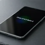 Fotografia macro de um smartphone premium com tela AMOLED exibindo a interface de atualização do sistema One UI 8.5 Beta 2, posicionado centralmente sobre uma superfície minimalista escura com iluminação cinematográfica.